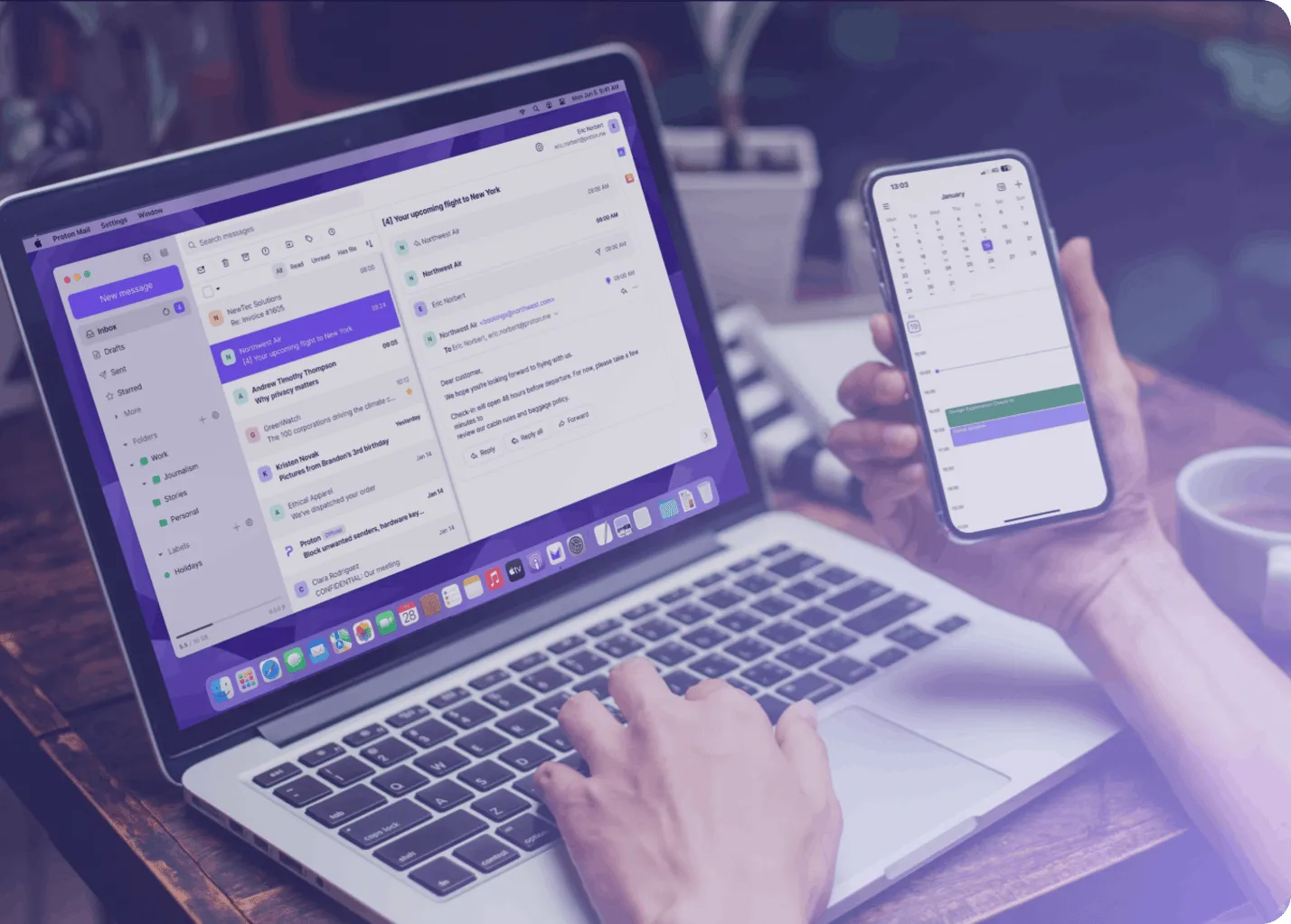 يد شخص تحمل هاتفًا يعرض تطبيق Proton Calendar، مع وجود جهاز كمبيوتر محمول على سطح المكتب يعرض تطبيق Proton Mail